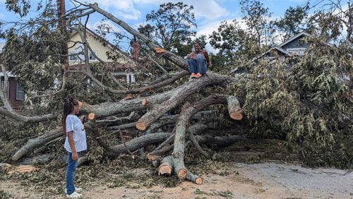 Qena Fabin, an Augusta resident, on top of the oak tree that toppled into her front yard because of Tropical Storm Helene while her seven-year-old daughter looks on. Fabin has to walk around the tree's massive roots or scramble over it to leave her yard on Sunday.