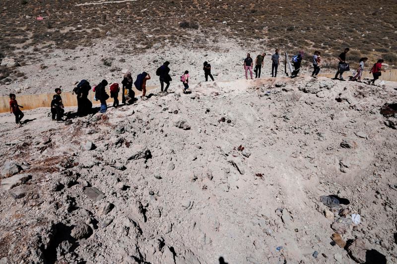 People carry their luggage as they cross into Syria on foot, through a crater caused by Israeli airstrikes aiming to block Beirut-Damascus highway at the Masnaa crossing, in the eastern Bekaa Valley, Lebanon, Saturday, Oct. 5, 2024. (AP Photo/Hassan Ammar)