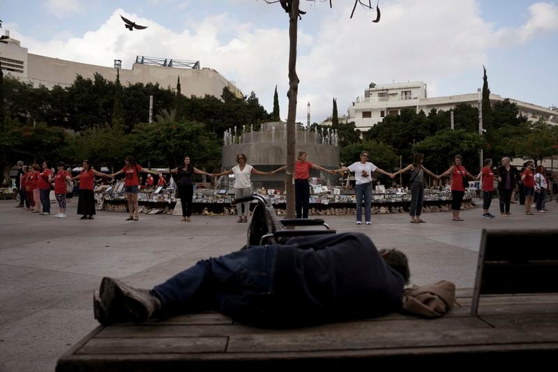 A man sleeps on a bench as people hold hands at a memorial marking the one-year anniversary of the Hamas attack on Israel, in Tel Aviv, Monday, Oct. 7, 2024. (AP Photo/Oded Balilty)