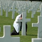 Dimitri Dodys took this picture during a visit to Normandy France in August 2017. The unidentified man was kneeling in prayer at this grave site. The Normandy American Cemetery and Memorial in France is located in Colleville-sur-Mer, on the site of the temporary American St. Laurent Cemetery, established by the U.S. First Army on June 8, 1944 as the first American cemetery on European soil in World War II. The cemetery site, at the north end of its half mile access road, covers 172.5 acres and contains the graves of 9,385 of American military dead, most of whom lost their lives in the D-Day landings and ensuing operations. On the Walls of the Missing, in a semicircular garden on the east side of the memorial, are inscribed 1,557 names. Rosettes mark the names of those since recovered and identified.