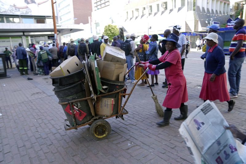 Street cleaners walk past people waiting in a bank queue in Harare, Zimbabwe, Thursday, April 25, 2024. Zimbabwe introduced the world's newest currency in April introduced the world's newest currency in April, the ZiG, or Zimbabwe Gold. (AP Photo/Tsvangirayi Mukwazhi)