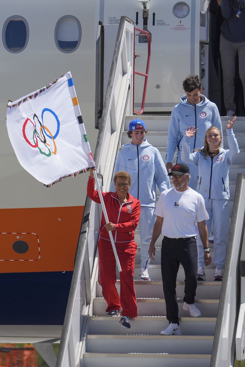 Los Angeles Mayor Karen Bass carrying the Olympic flag, LA28 chairman Casey Wasserman, front right, and Team USA Olympians, skateboarder Tate Carew, second from left, diver Delaney Schnell, rear right, and volleyball player Micah Ma'a, top right, arrive at Los Angeles International Airport, Monday, Aug. 12, 2024. (AP Photo/Damian Dovarganes)