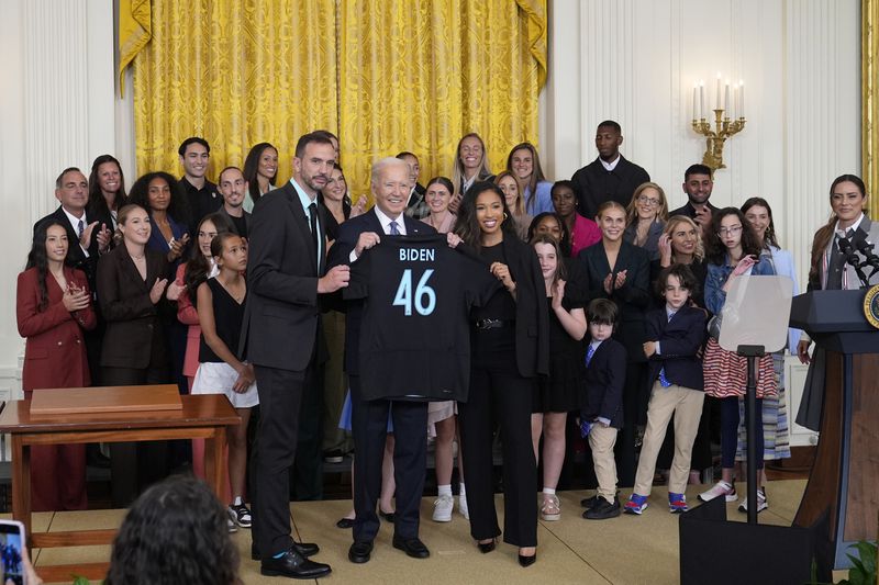 President Joe Biden, center, holds a team jersey from NJ/NY Gotham FC that was presented to him by head coach Juan Carlos Amoros, left, and teammate Midge Purse, right, during an event in the East Room of the White House in Washington, Monday, Sept. 23, 2024, to welcome the NJ/NY Gotham FC and celebrate their 2023 NWSL championship. (AP Photo/Susan Walsh)