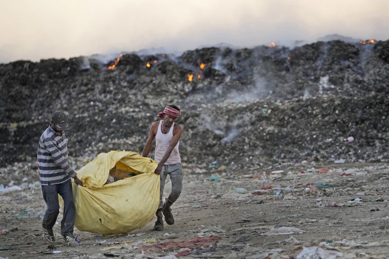 Usmaan Shekh, right, carries, with help, a bag of recyclable material collected from a garbage dump site during a heat wave on the outskirts of Jammu, India, Wednesday, June 19, 2024. Shekh and his family are among millions of people who scratch out a living searching through India's waste — and climate change is making a hazardous job more dangerous than ever.(AP Photo/Channi Anand)