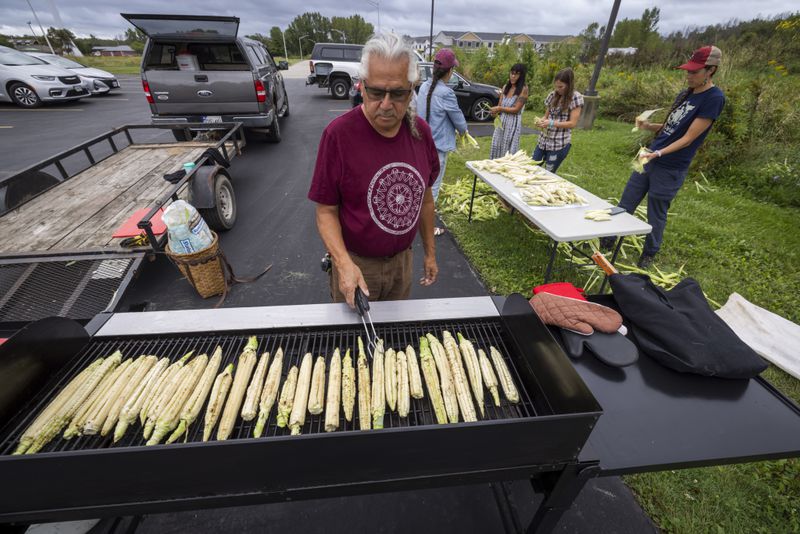 Robin John roasts cobs of corn as other members of Ohe·laku, a non-profit that works with the families planting crops, shuck cobs of corn before they are roasted, cut and bagged on the Oneida Indian Reservation, Friday, Aug. 30, 2024, in Oneida, Wis. (AP Photo/Mike Roemer)