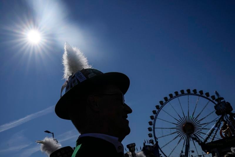 A man with a traditional Bavarian hat is pictured in the sun at start of the 189th 'Oktoberfest' beer festival in front of paintings showing Munich landmarks in Munich, Germany, Saturday, Sept. 21, 2024. (AP Photo/Matthias Schrader)