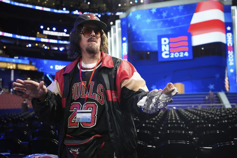 Stephen Colbert, host of the "Late Show," seen in character, eats a hotdog as he walks on the convention floor before the Democratic National Convention Sunday, Aug. 18, 2024, in Chicago. (AP Photo/Paul Sancya)