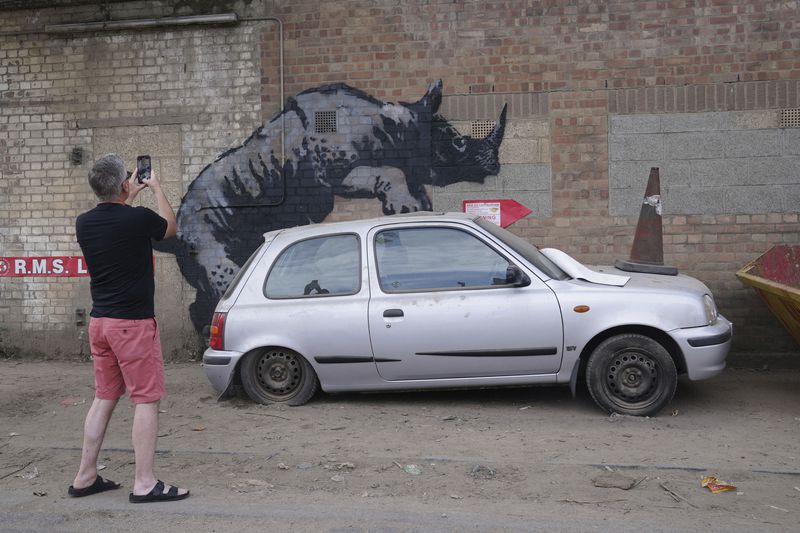 A man photographs a new artwork unveiled by Banksy, depicting a rhinoceros which looks as though it is climbing on top of a car, the eighth artwork in his animal-themed collection, on Westmoor Street in Charlton, south east London, Monday, Aug. 12, 2024. (Lucy North/PA via AP)