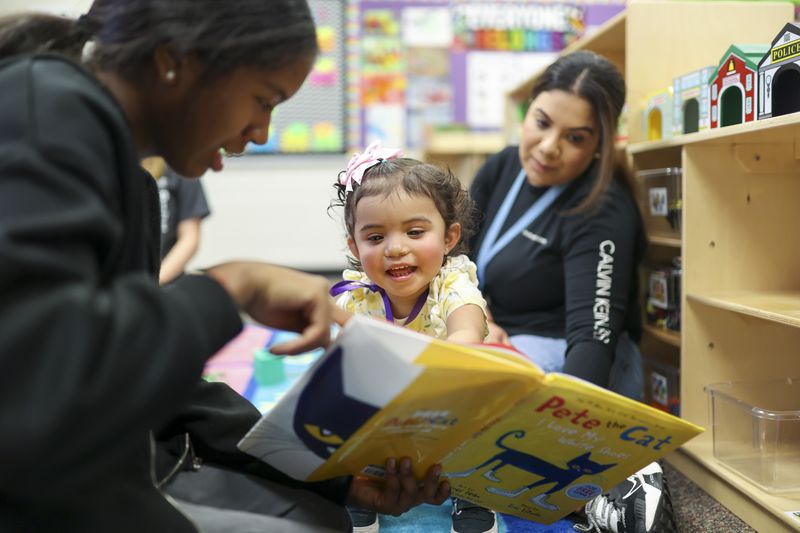 Eighth grade student Mia Avouwadan reads the book "Pete the Cat" to 20-month-old Daniela Trevino at the Play 2 Learn program at Duluth Middle School, Wednesday, Nov. 8, 2023. Daniela’s mother Ilse Trevino is shown at right. These students are part of the teaching profession program and have an interest in a career in education. (Jason Getz / Jason.Getz@ajc.com)