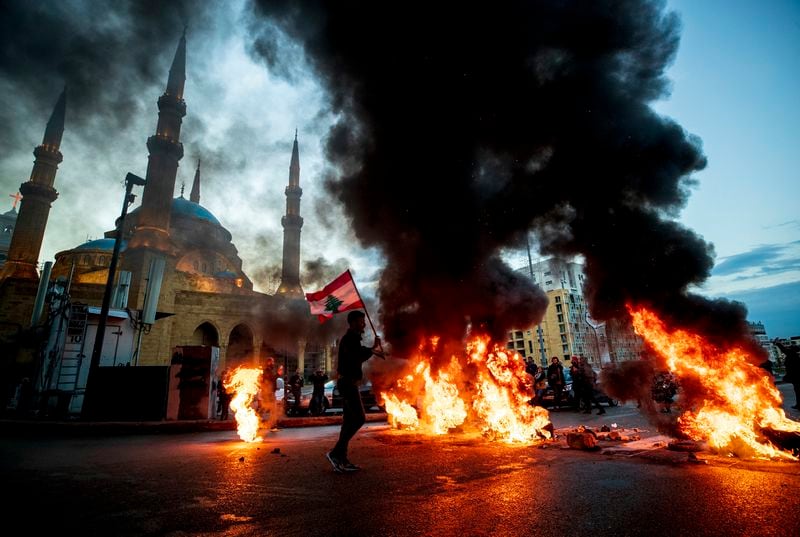 FILE - A protester holds up a Lebanese national flag as he walks in front of burning tires that are blocking a main road, during a protest in downtown Beirut, on March 3, 2021. (AP Photo/Hassan Ammar, File)