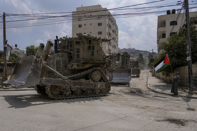 A man waves the Palestinian flag as a convoy of Israeli military bulldozers drive by during an army raid in Jenin, West Bank, Monday, Sept. 2, 2024. (AP Photo/Majdi Mohammed)