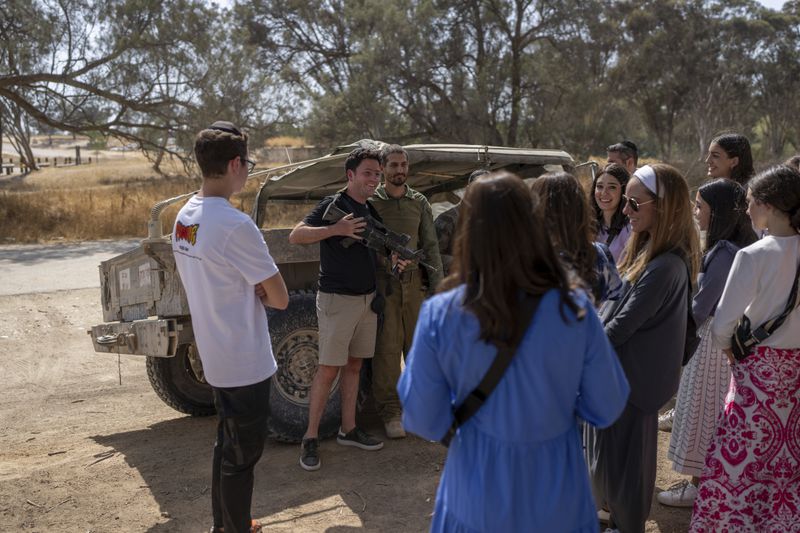 An Israeli reservist poses for a picture with a tourist from Mexico, who is holding the soldier's M16 rifle, at the site of the Tribe of Nova music festival, where at least 364 people were killed and abducted during the Hamas attack on Oct. 7, near Kibbutz Re'im, southern Israel on Thursday, June 20, 2024. A new kind of tourism has emerged in Israel in the months since Hamas’ Oct. 7 attack. For celebrities, politicians, influencers and others, no trip is complete without a somber visit to the devastated south that absorbed the brunt of the assault near the border with Gaza. (AP Photo/Ohad Zwigenberg)