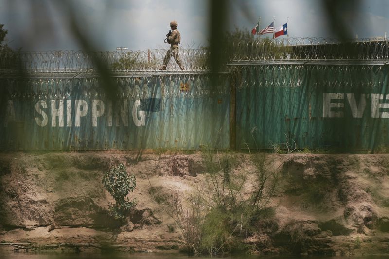 A member of the Texas Army National Guard patrols the Rio Grande from atop a line of shipping containers in Eagle Pass, Texas, on Friday, Aug. 9, 2024. (Jordan Vonderhaar/The Texas Tribune via AP)