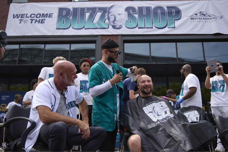 Seattle Mariners barber Mark Basinger puts the finishing touches on catcher Cal Raleigh's shaved head with former Mariners player Jay Buhner, left, watching, on Buhner Buzz Night, Thursday, June 13, 2024, in Seattle. The promotion is based on Buhner's shaved-head style. (AP Photo/John Froschauer)
