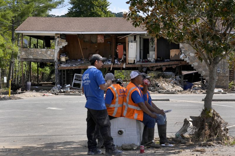 Workers helping with clean up efforts stop for lunch under the shade of a tree as a building destroyed by Hurricane Helene is seen in the background Saturday, Oct. 5, 2024, in Newport, Tenn. (AP Photo/Jeff Roberson)