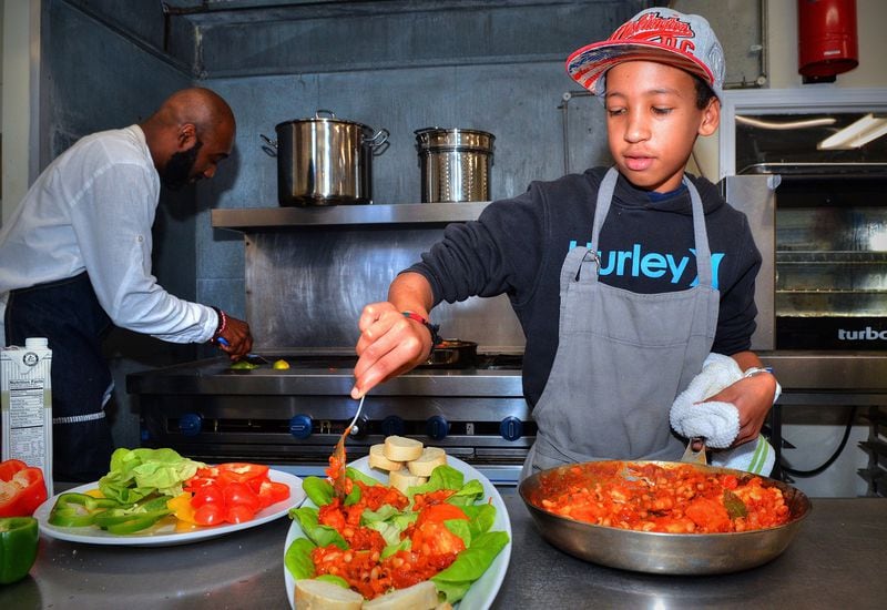 Chef Cheikh Ndiaye (background) guides 12-year-old Fallou Diouf’s cooking efforts. Here Fallou, whose father is Senegalese, does the final touches for the Ndambe (black-eyed pea ragout served on toast or in lettuce wraps). CONTRIBUTED BY CHRIS HUNT PHOTOGRAPHY