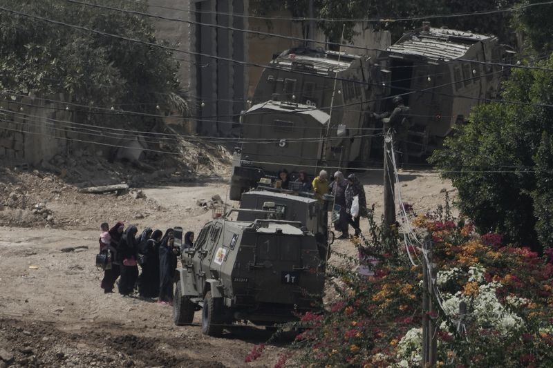 Palestinians stand in line next to Israeli armoured vehicles during a military operation in the West Bank Jenin refugee camp, Saturday, Aug. 31, 2024. (AP Photo/Majdi Mohammed)