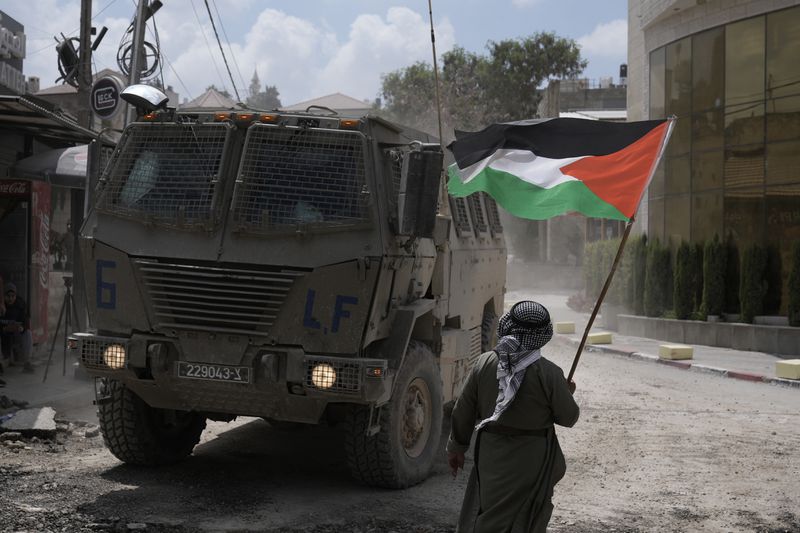 A man waves a Palestinian flag as an Israeli armoured vehicle moves on a street during a military operation in the West Bank refugee camp of Nur Shams, Tulkarem, Thursday, Aug. 29, 2024. (AP Photo/Majdi Mohammed)