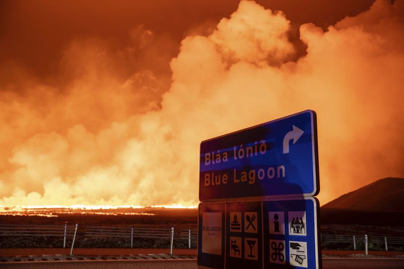 The sign with the direction to the blue lagoon with the new eruption in the background in Grindavik, Iceland, Thursday, Aug. 22, 2024, (AP Photo/Marco di Marco)