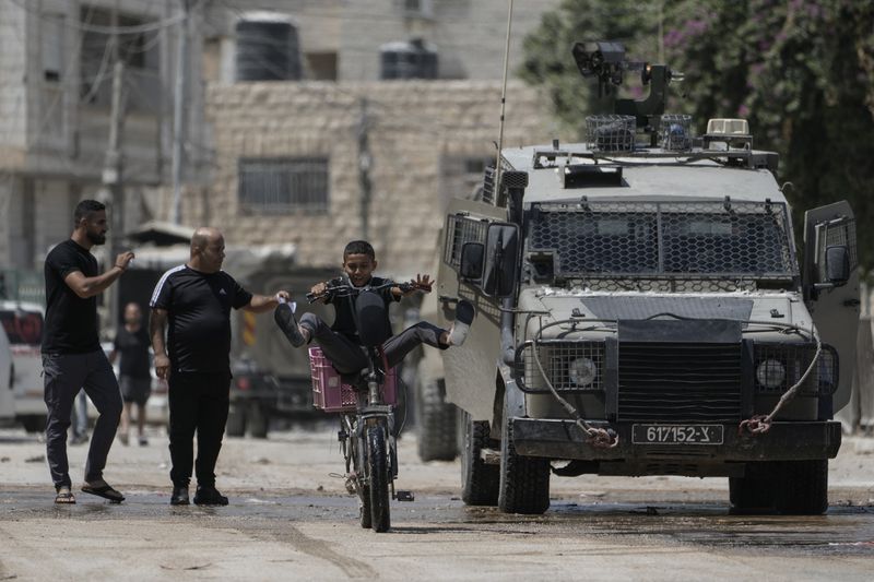 A youth rides his bicycle past an Israeli armoured vehicle during a military operation in the West Bank city of Jenin, Wednesday, Aug. 28, 2024. (AP Photo/Majdi Mohammed)