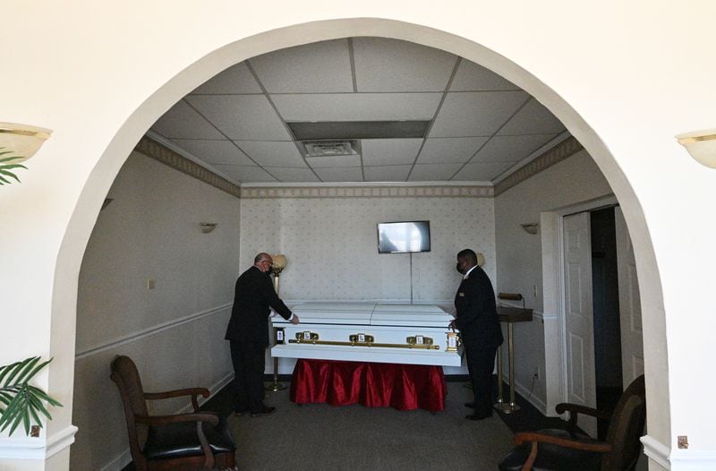 Garry Duty, left, and Deric Thomas, both funeral home staff, place a casket in a viewing room at Dudley Funeral Home in Dublin in March. In August and September of 2020, the funeral home was handling two or three services a day. It usually does five or six a week. (Hyosub Shin / Hyosub.Shin@ajc.com)
