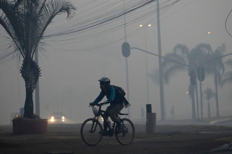 A cyclist pedals through the smoke in Brasilia, Brazil, coming from a fire in the environmentally protected area of Brasilia National Park, early Monday, Sept. 16, 2024. The head of the agency that manages protected areas, Mauro Pires, told the local press that the fire is man-made and appears to have started near the edge of a farm. (AP Photo/Eraldo Peres)