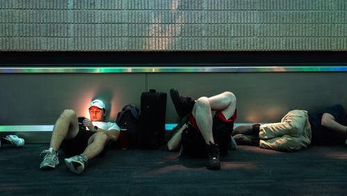 Brian Hauk of Detroit (left) looks at his phone while other stranded travelers sleep at the Hartsfield-Jackson airport domestic terminal late in the evening in Atlanta on Saturday, July 20, following a global technology outage that has hampered airlines and other industries. (Arvin Temkar / AJC)