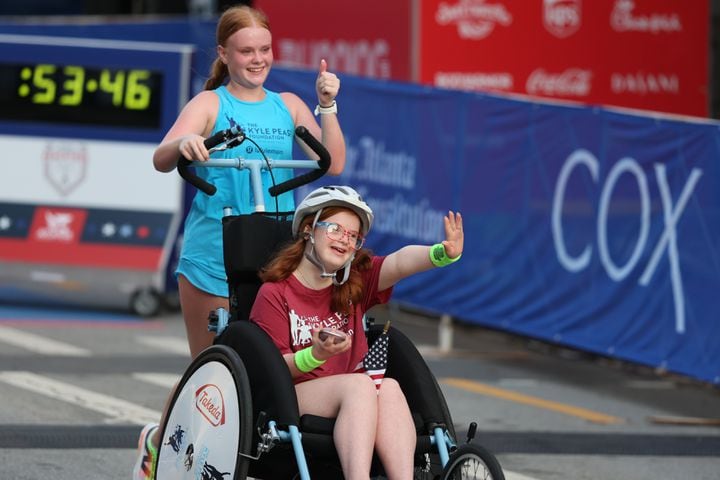 Darden Glass being pushed by sister Anna Glass crosses the finish line at the 55th running of the Atlanta Journal-Constitution Peachtree Road Race in Atlanta on Thursday, July 4, 2024.   (Jason Getz / AJC)
