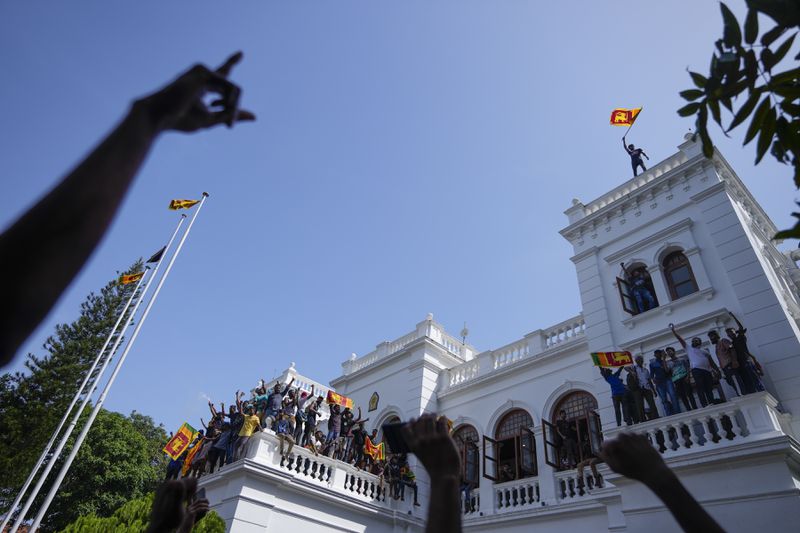 A Sri Lankan protester waves the national flag from the rooftop of Sri Lankan Prime Minister Ranil Wickremesinghe's office, demanding he resign after president Gotabaya Rajapaksa fled the country amid economic crisis in Colombo, Sri Lanka, Wednesday, July 13, 2022. (AP Photo/Eranga Jayawardena, File)