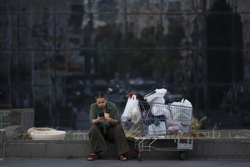 A woman looks at her phone while eating a sandwich next to a shopping cart in Haifa, Israel, Thursday, Aug. 15, 2024. Israel's economy is suffering from the nearly 11-month war with Hamas, as its leaders grind ahead with its offensive in Gaza that threatens to escalate into a wider conflict. (AP Photo/Leo Correa)