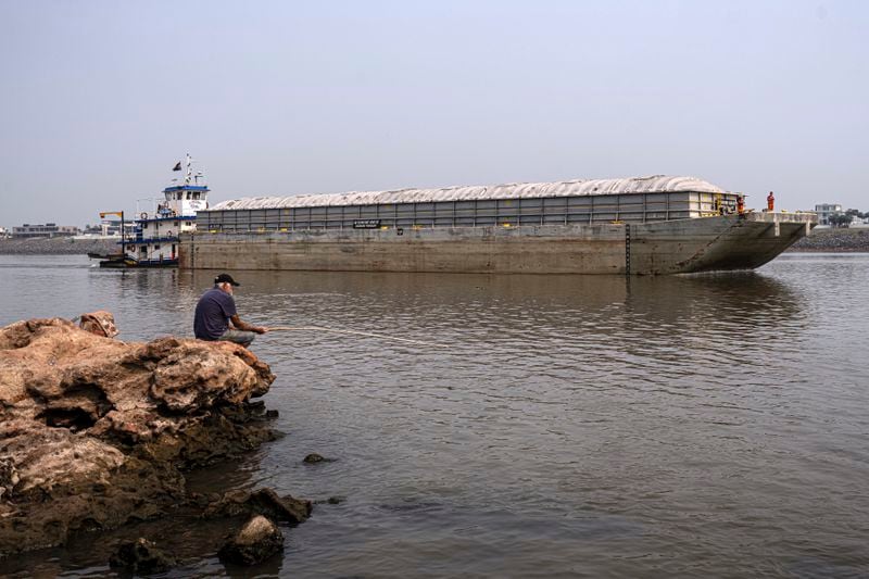 A man fishes on the shore of the Paraguay River where a tugboat pushes a barge in Mariano R. Alonso, Paraguay, Monday, Sept. 9, 2024. Water levels have plunged to their lowest-ever level amid a drought, according to Paraguay's Meteorology and Hydrology Office. (AP Photo/Jorge Saenz)