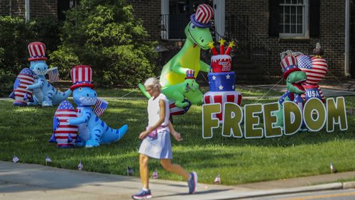Alyson Cohen strolled by the display of dinosaurs on Dunwoody Club Drive near Braffington Court in Dunwoody on Friday, June 9, 2023. The display of dinosaurs first appeared in the neighborhood in 2020 then quickly evolved into holiday themed displays along with encouraging messages.  (John Spink / John.Spink@ajc.com)