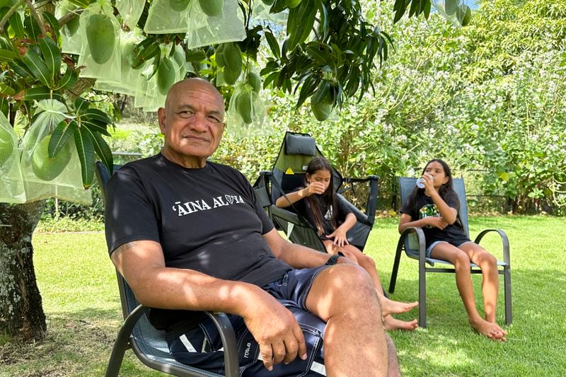 Randy Awo sits in his backyard with his granddaughters in Wailuku, Hawaii, on Thursday, July 18, 2024. As a Native Hawaiian Maui resident and a retired administrator in the state Department of Land and Natural Resources, Awo said he is concerned about storing debris from last year's deadly fire anywhere on the island. (AP Photo/Jennifer Sinco Kelleher)