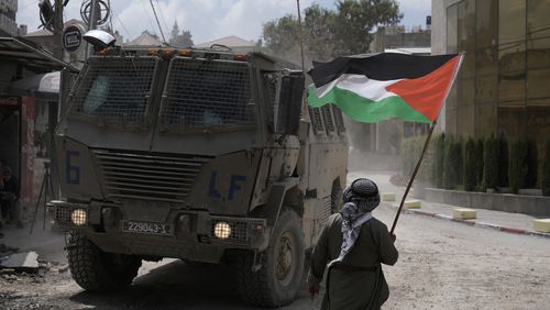 A man waves a Palestinian flag as an Israeli armoured vehicle moves on a street during a military operation in the West Bank refugee camp of Nur Shams, Tulkarem, Thursday, Aug. 29, 2024. (AP Photo/Majdi Mohammed)