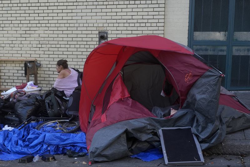 A person sits behind a tent on a sidewalk in San Francisco, Thursday, Aug. 29, 2024. (AP Photo/Jeff Chiu)