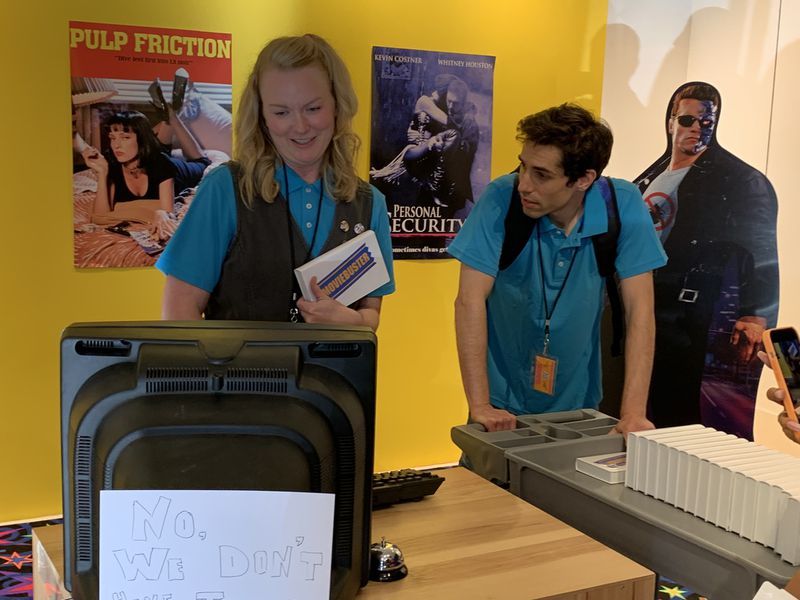 The "Save the Video Store" interactive at The Avenue West Cobb is a faux video rental store from the 1990s. Actors Erin Stegeman (left, who created the experience) and Zach Canner play employees. RODNEY HO/rho@ajc.com