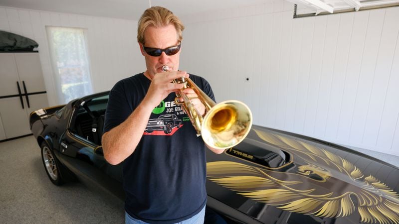 Musician Joe Gransden plays his trumpet next to his meticulously restored 1979 Pontiac Trans Am in his home’s garage. 
(Miguel Martinez / AJC)