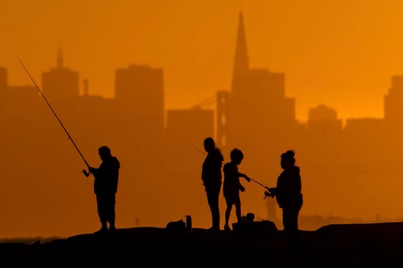With the San Francisco skyline behind them, people fish off a jetty Monday, July 1, 2024, in Alameda, Calif. An extended heatwave predicted to blanket Northern California has resulted in red flag fire warnings and the possibility of power shutoffs beginning Tuesday. (AP Photo/Noah Berger)