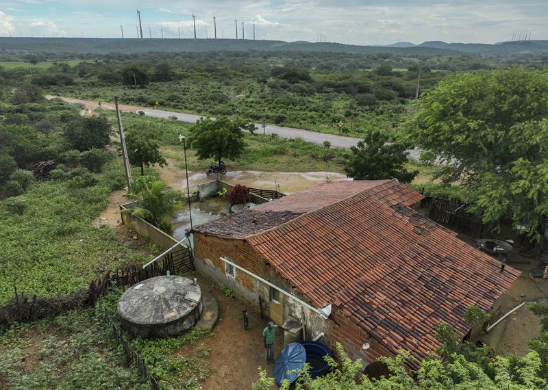 Joao de Souza Silva stands in his backyard, with wind turbines visible in the distance in Sumidouro, Piaui state, Brazil, Wednesday, March 13, 2024. Wind energy is booming in Brazil's Northeast, but some projects are drawing criticism as it becomes clear that certain communities have benefited while others have not. (AP Photo/Andre Penner)
