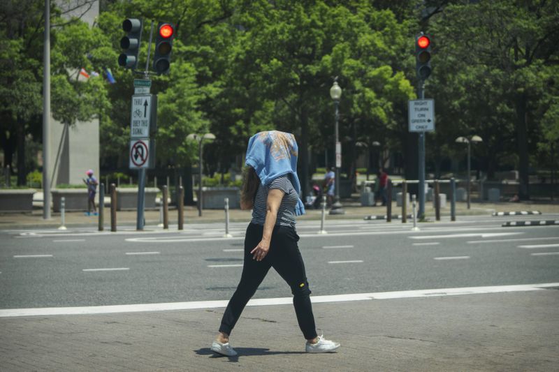 A woman wearing a sweatshirt on her head to protect herself from the sun walks across a crosswalk in downtown Washington, Wednesday, June 19, 2024. High temperatures are expected to stay in the 90s in the nation's capital for the rest of the week as a heat wave builds into the Northeast. (AP Photo/Mark Schiefelbein)