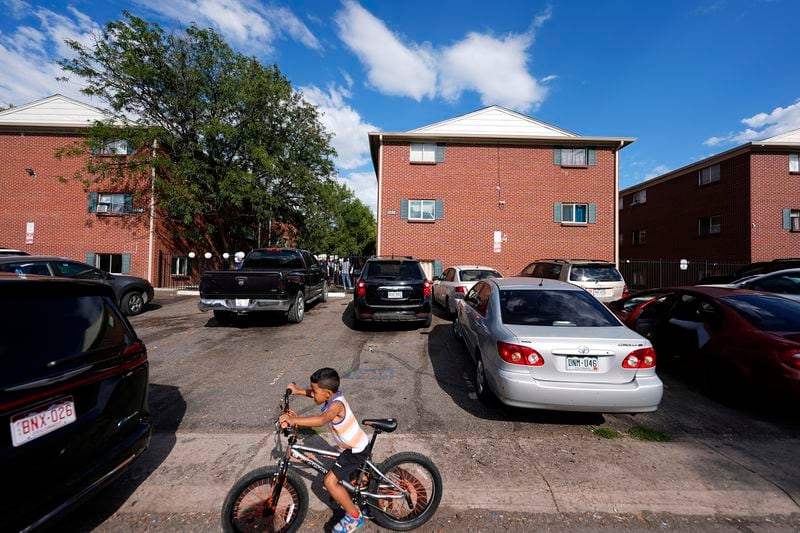 A boy guides his bicycle past apartment buildings as a rally staged by the East Colfax Community Collective is held in the courtyard to address chronic problems in the apartment buildings occupied by people displaced from their home countries in central and South America Tuesday, Sept. 3, 2024, in Aurora, Colo. (AP Photo/David Zalubowski)