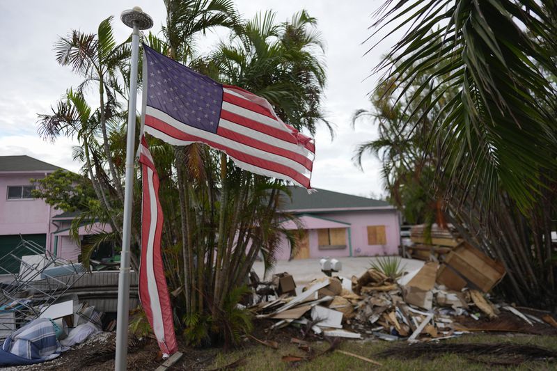 A tattered American flag flaps outside a home as furniture and household items damaged by Hurricane Helene flooding sit piled along the street awaiting pickup, ahead of the arrival of Hurricane Milton, in Holmes Beach on Anna Maria Island, Fla., Tuesday, Oct. 8, 2024. (AP Photo/Rebecca Blackwell)