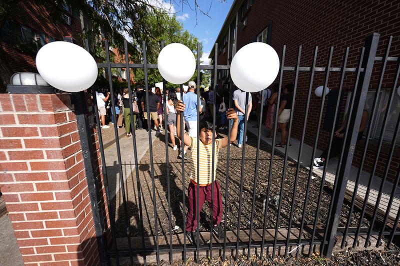 A young boy climbs an iron fence to reach balloons during a rally staged by the East Colfax Community Collective to address chronic problems in the apartment buildings occupied by people displaced from their home countries in central and South America Tuesday, Sept. 3, 2024, in Aurora, Colo. (AP Photo/David Zalubowski)