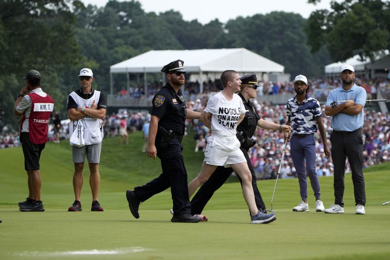 One of several protesters is led away after they ran onto the course on at the 18th hole as Akshay Bhatia, second from right, and Scottie Scheffler, right, watch during the final round of the Travelers Championship golf tournament at TPC River Highlands, Sunday, June 23, 2024, in Cromwell, Conn. (AP Photo/Seth Wenig)