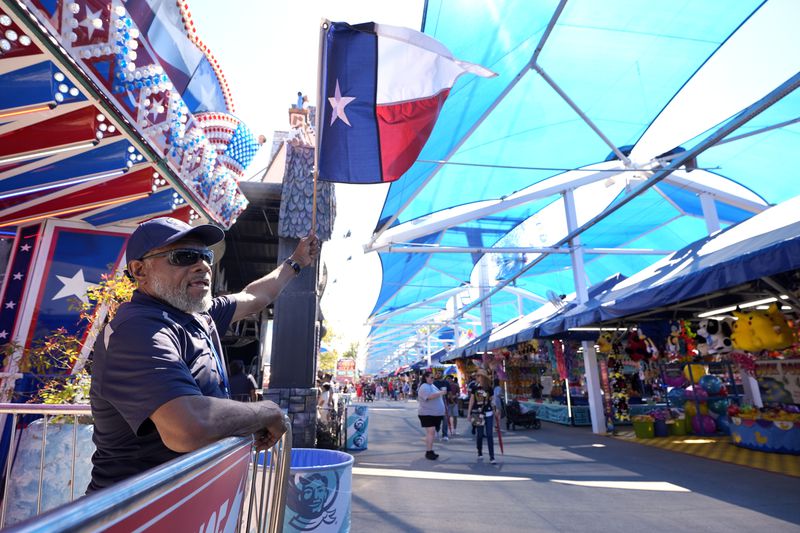 J.R., waves a Texas state flag as he attempts to attract guests to his game attraction at the State Fair of Texas in Dallas, Friday, Sept. 27, 2024. (AP Photo/Tony Gutierrez)