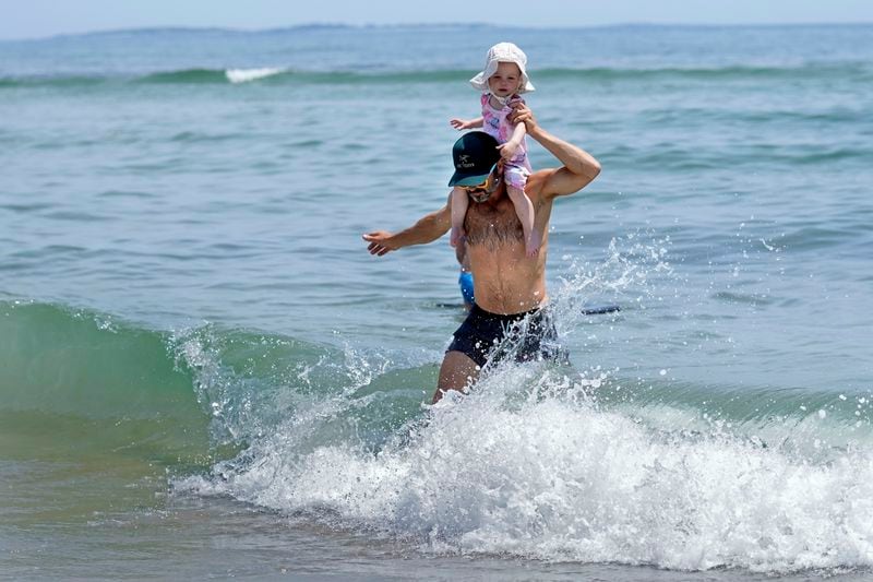 Jerome Quirion of Sherbrooke, Quebec, Canada, wades back to shore after venturing into the chilly Atlantic Ocean with his 18-month-old daughter, Amelie, while vacationing, in Old Orchard Beach, Maine, Tuesday, June 18, 2024. The heat wave that has been hitting much of the United States is now moving into the Northeast. (AP Photo/Robert F. Bukaty)
