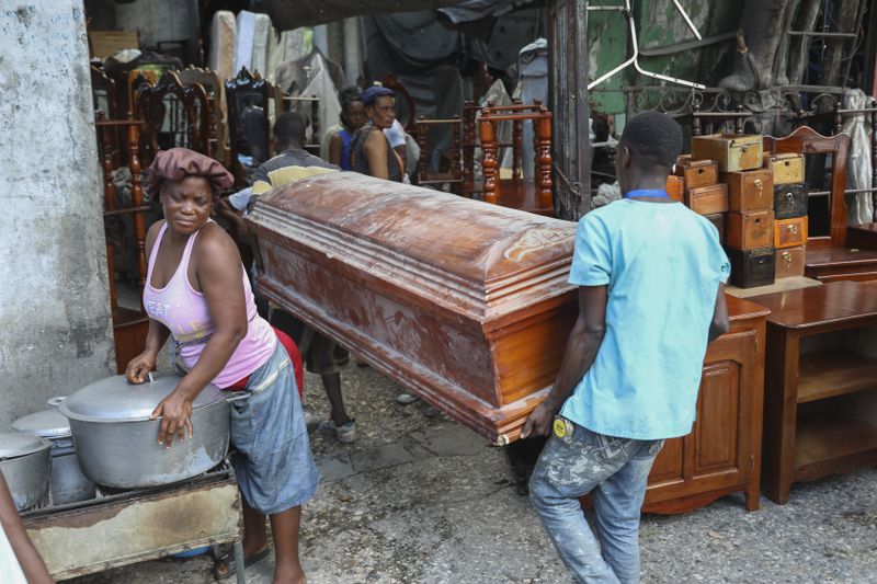 People carry an empty coffin past a street food vendor into the shop that is buying it to repair and sell in Port-au-Prince, Haiti, Friday, Sept. 13, 2024. (AP Photo/Odelyn Joseph)