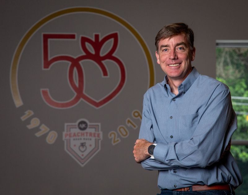 Atlanta track club’s Executive Director Rich Kenah in front of the logo for the 50th Running of the AJC Peachtree Road Race in the track club’s office June 4, 2019. STEVE SCHAEFER / SPECIAL TO THE AJC