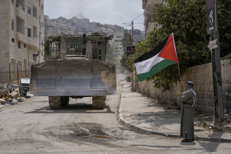 A man waves the Palestinian flag as a convoy of Israeli military bulldozers drive by during an army raid in Jenin, West Bank, Monday, Sept. 2, 2024. (AP Photo/Majdi Mohammed)