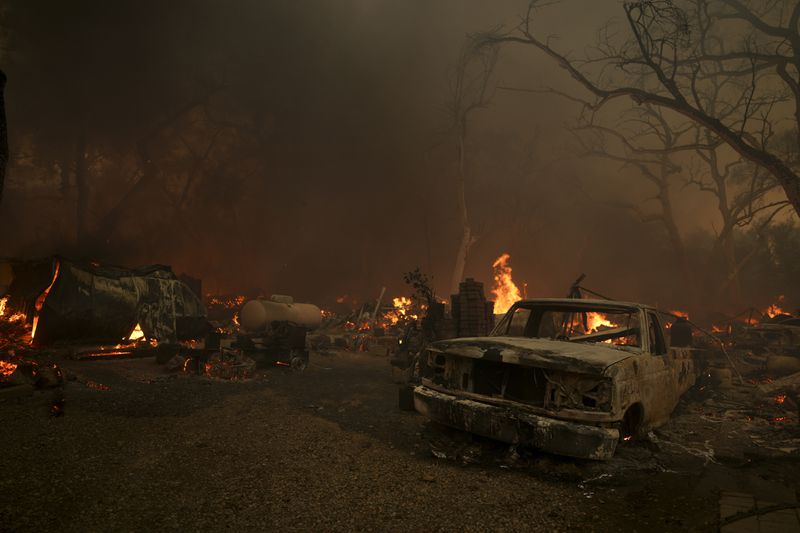 A destroyed car sits as structures burn after the Airport Fire swept through Tuesday, Sept. 10, 2024, in El Cariso, an unincorporated community in Riverside County, Calif. (AP Photo/Eric Thayer)